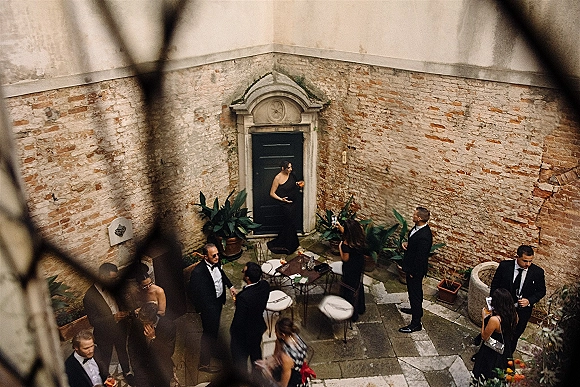 Wedding cocktail hour with guests mingling around a bistro table, holding cocktails on a stone patio by a brick wall and black door, with orange flowers