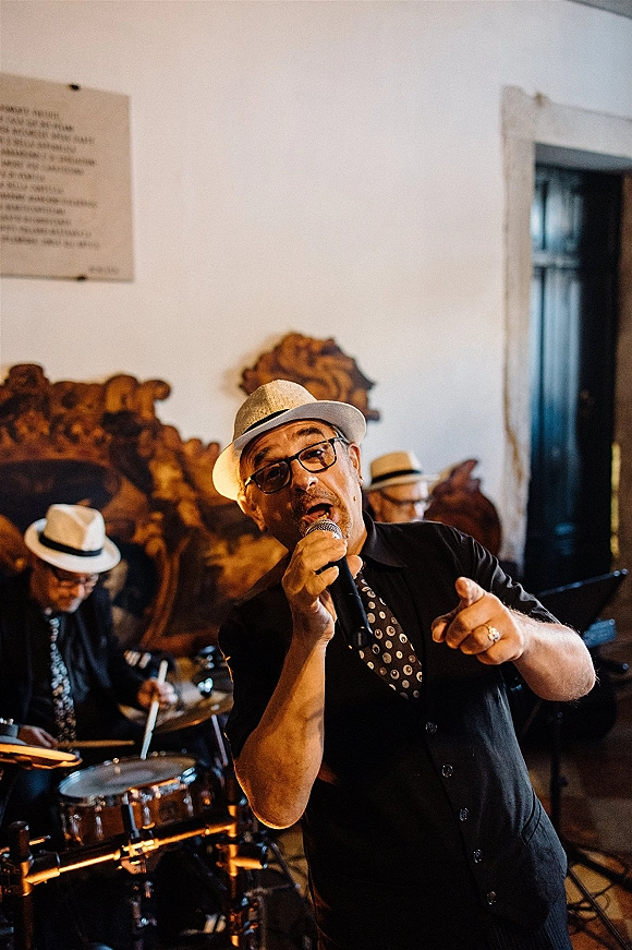 Wedding band performance with singer at a microphone in fedora and glasses, drummer behind at drum kit on an indoor stage area