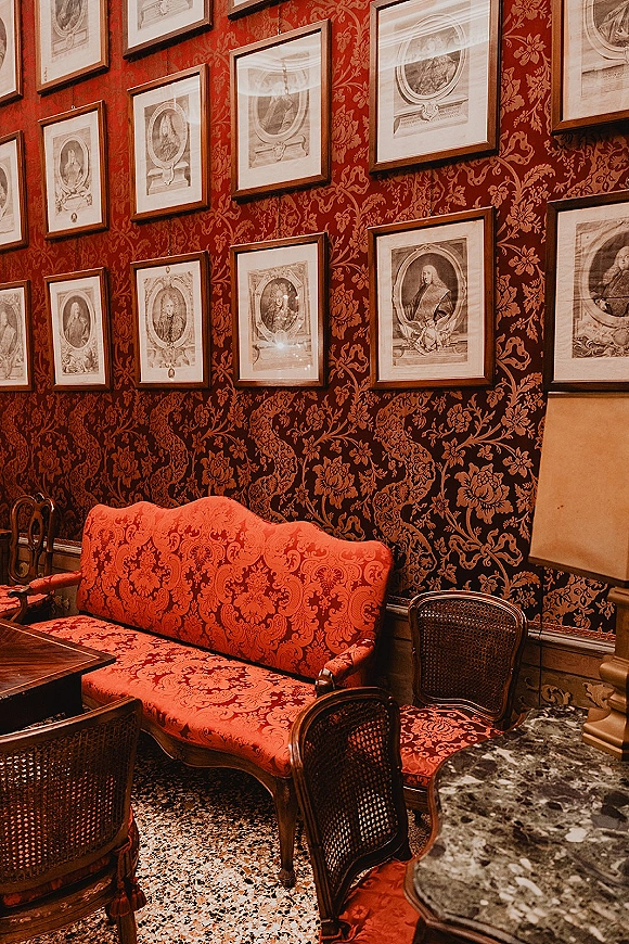 Vintage lounge seating with a red upholstered sofa and cane bistro chairs around a marble table, framed portraits on red damask walls