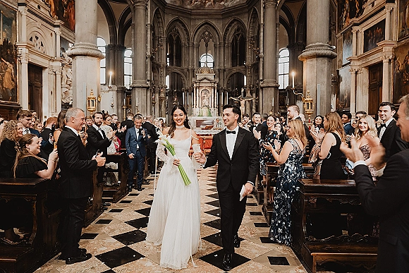 Wedding recessional as bride and groom walk the aisle hand in hand, calla lily bouquet and tuxedo amid pews in a grand church interior