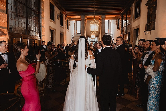 Wedding reception entrance as newlyweds walk in from behind, veil and tuxedo framed by cheering guests with champagne in a chandelier-lit hallway
