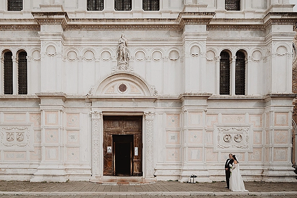 Couple portrait of bride holding bouquet beside groom in black suit at church entrance, her wedding dress and veil framed by arched doors