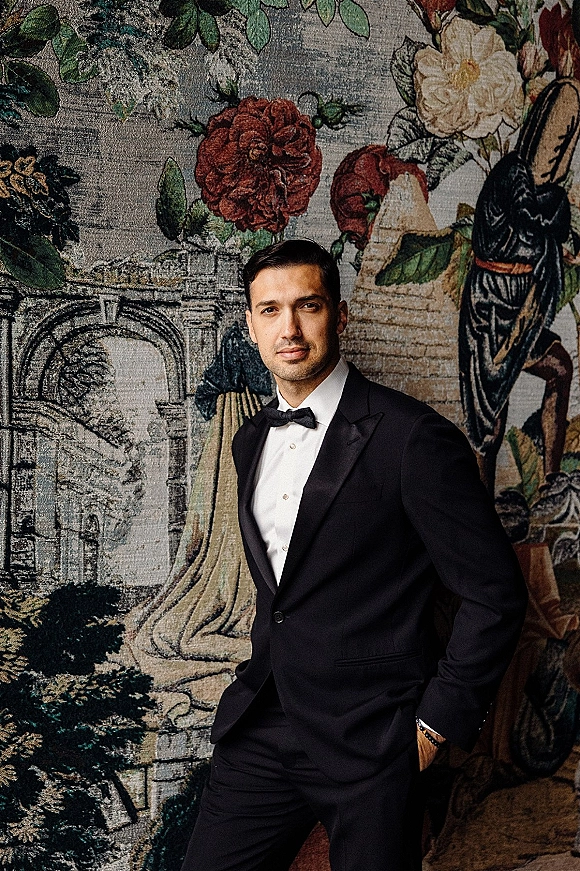 Groom portrait in a black tuxedo with bow tie and wristwatch, standing indoors against a vintage floral tapestry wall hanging
