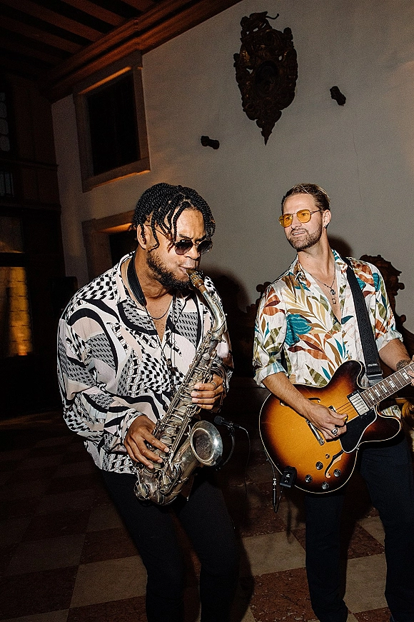 Wedding band performance with saxophone and electric guitar as musicians play indoors beneath wood ceiling beams and ornate wall decor