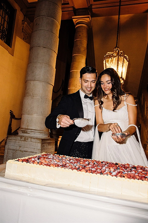 Wedding cake moment as bride and groom sift powdered sugar over a berry-topped sheet cake on a dessert table in a warm stone hallway