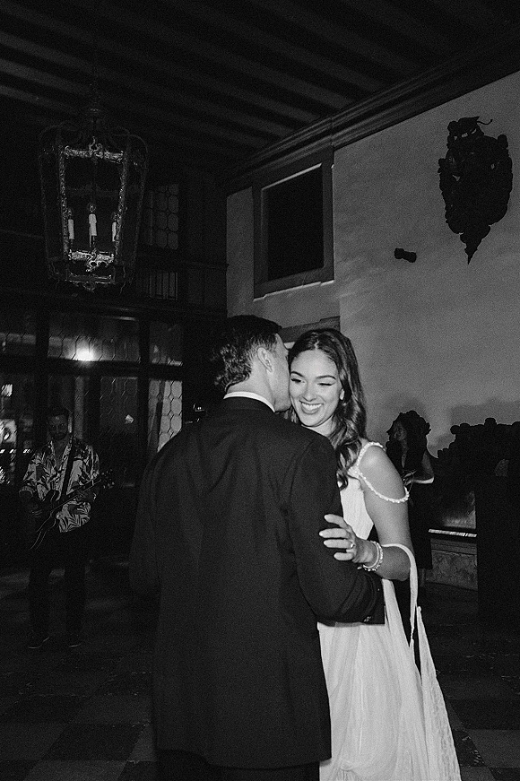 First dance as bride in pearl strap wedding dress slow dances with groom in a black suit under chandelier lanterns at reception