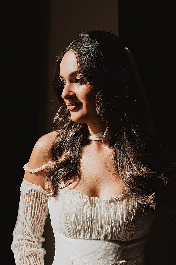 Bridal portrait of a bride looking away in window light, wearing an off-the-shoulder lace-sleeved gown and choker in a dark interior