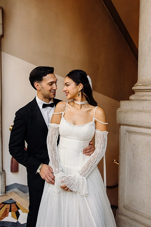 Couple portrait of bride and groom embrace on a stairwell, her veil and off-the-shoulder long-sleeve gown beside his black tuxedo