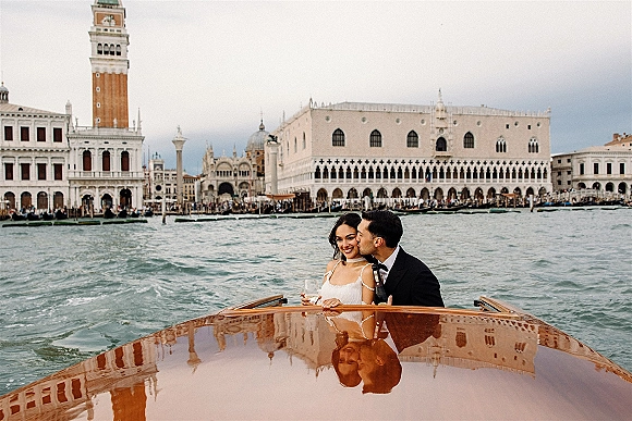 Couple portrait on a wedding boat portrait, groom kissing bride’s cheek as she holds champagne, with canal and bell tower behind