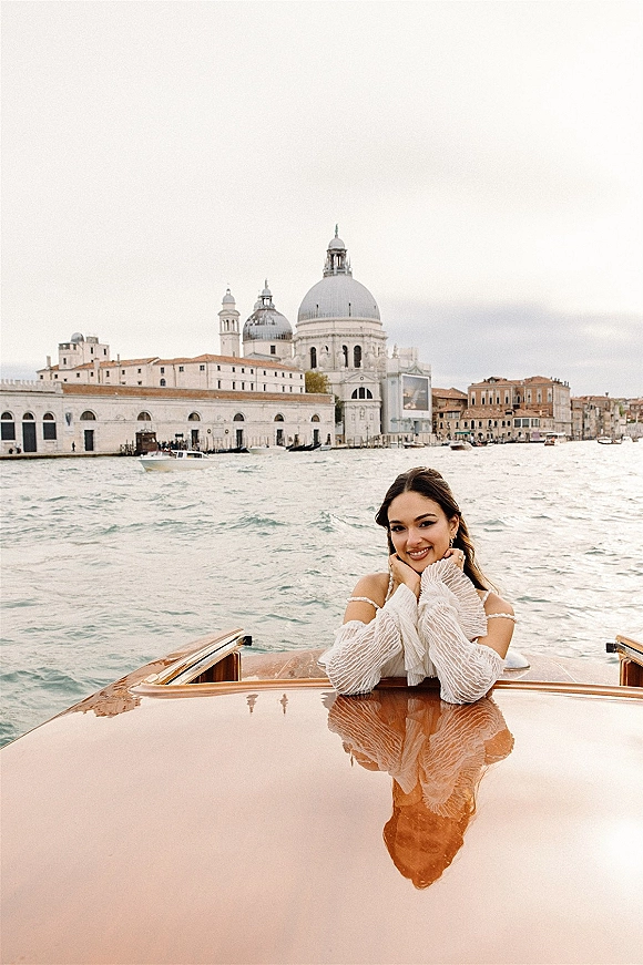Bridal portrait of a bride on boat in an off the shoulder long-sleeve dress, leaning by water with historic buildings and cloudy sky behind