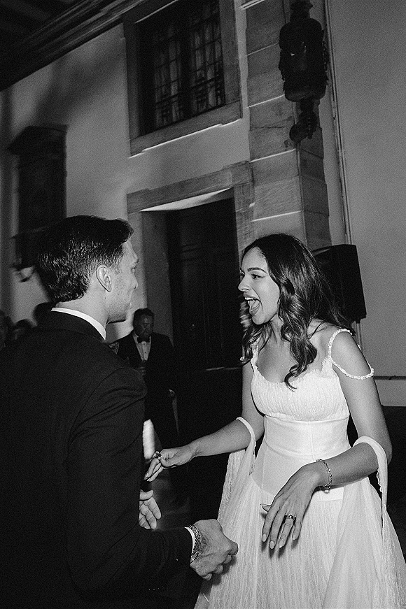 First dance moment as bride laughs holding the groom’s hand, off-the-shoulder gown and rings visible in a black-and-white reception hall
