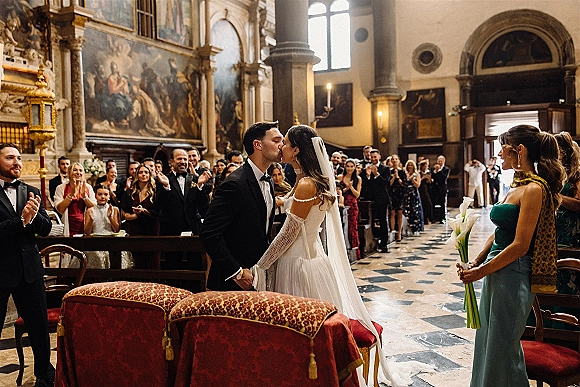 Wedding kiss in a church as the bride in an off-the-shoulder dress and veil leans into the groom in a tux, guests lining the aisle