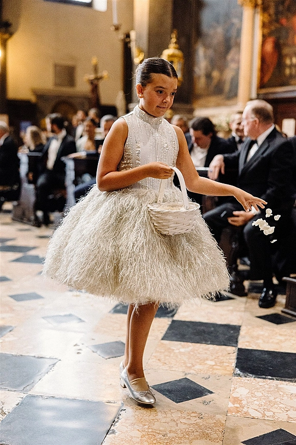 Flower girl tossing petals holds a basket in a sparkly tulle dress with pearl studs and silver shoes on a church aisle
