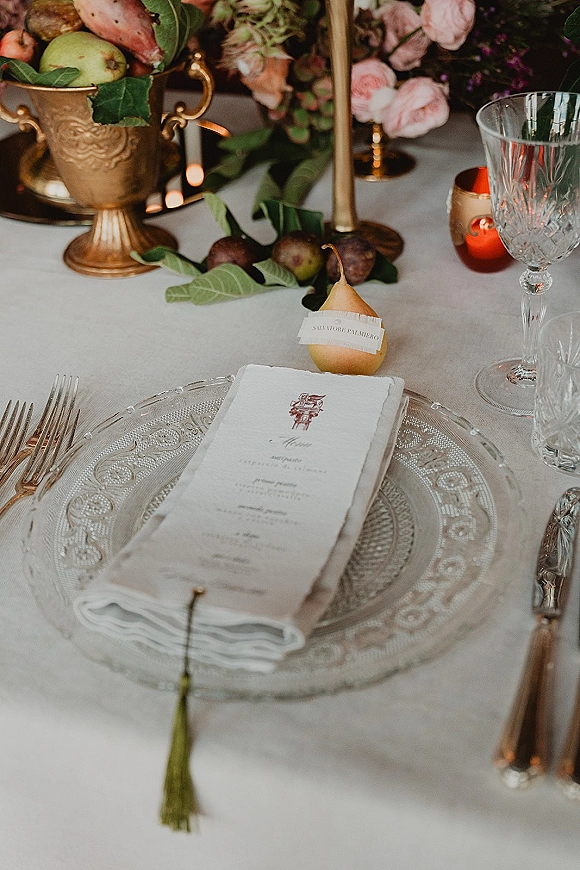 Wedding tablescape with a wedding place setting featuring a pear place card, blush roses, greenery garland, and gold taper candles on white linen