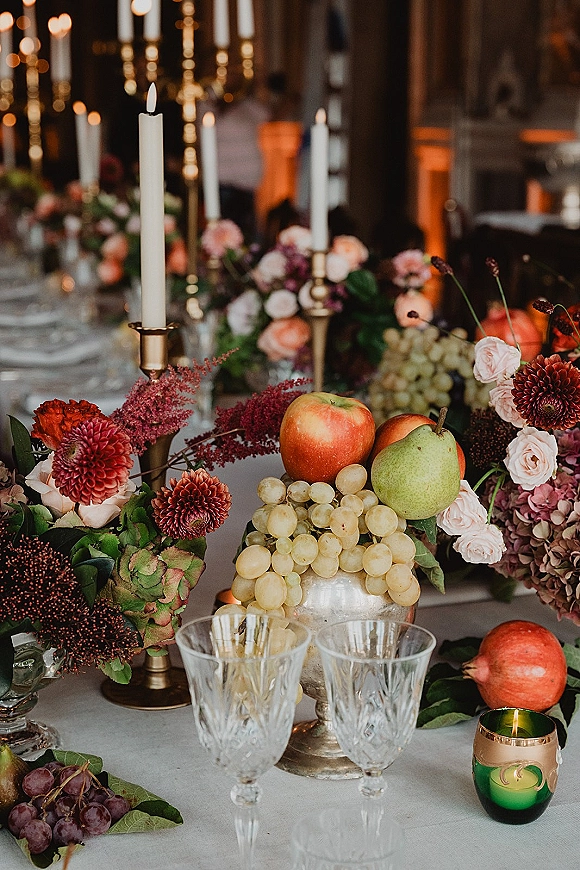 Reception tablescape wedding table centerpiece with taper candles in brass holders, lush florals, fruit, and crystal goblets in a dim room