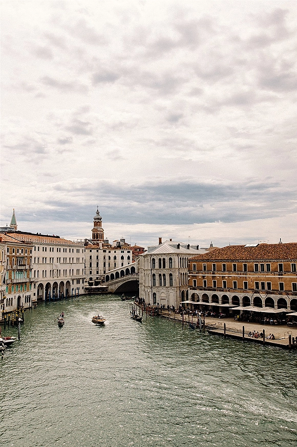 Canal cityscape with historic canal architecture, boats gliding under an arched bridge past waterfront buildings and cafe umbrellas under cloudy sky
