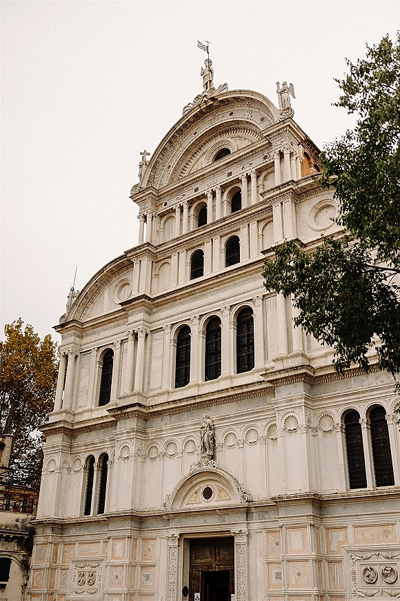 Church exterior with ornate stonework and statues, featuring arched windows, columns, and a cross above wooden doors against trees and sky