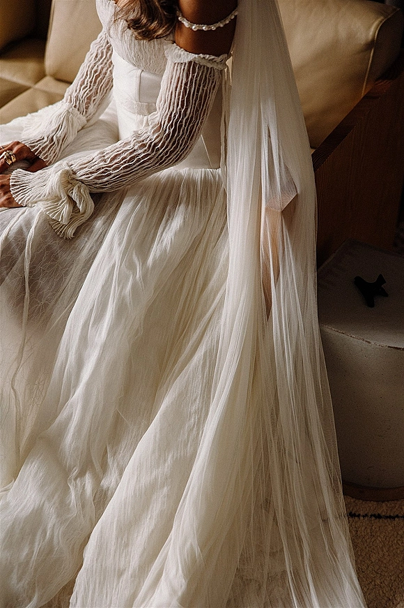 Wedding dress close-up with sheer sleeve wedding dress details, sash belt, pearl necklace and veil as bride sits on a beige chair indoors
