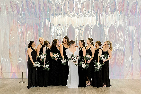 Bridesmaids group photo with bride centered, bridesmaids in black dresses holding bouquets, posing by an iridescent wall backdrop indoors
