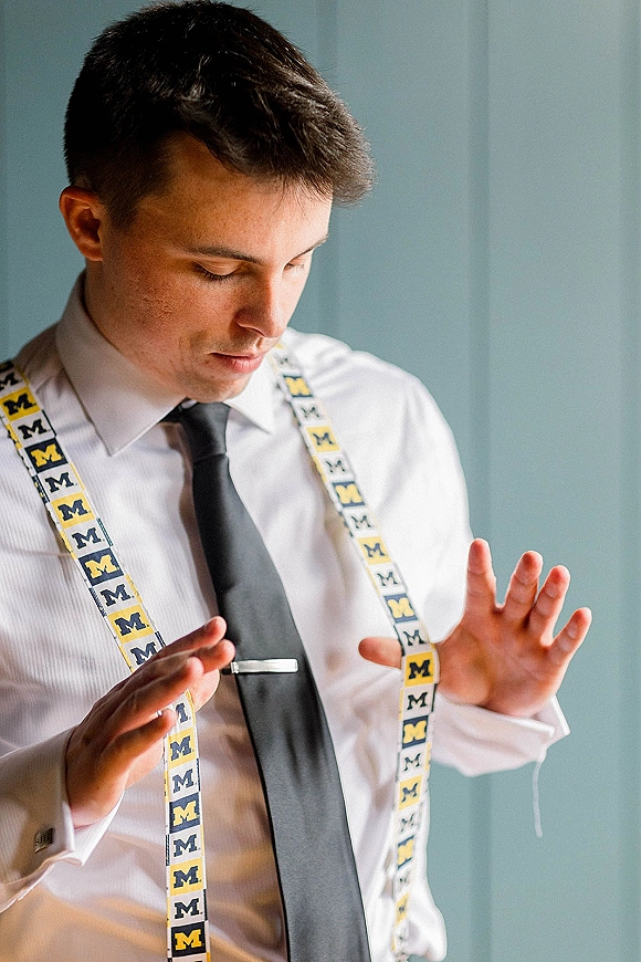 Groom getting ready adjusting groom suspenders over a white dress shirt, fastening a tie clip by an indoor wall before the ceremony