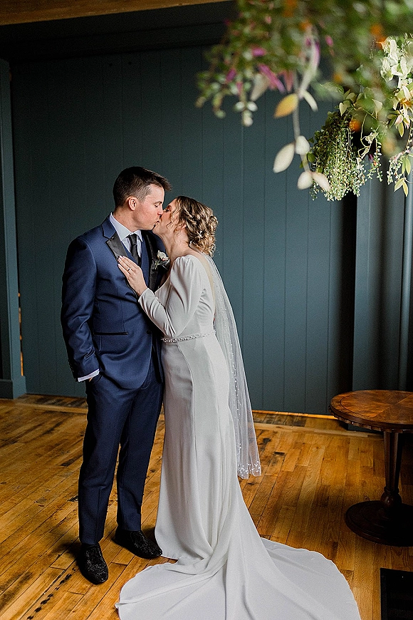 Wedding kiss portrait of bride and groom kissing, bride’s hand on his chest beneath hanging greenery against a dark paneled wall indoors