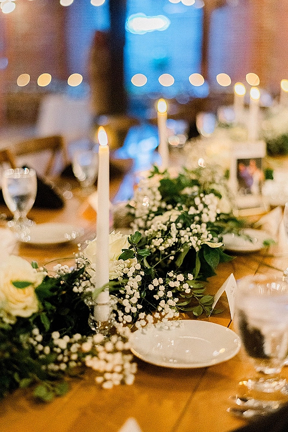Reception tablescape with greenery garland centerpiece, white taper candles, roses, place cards and glassware under warm string lights indoors