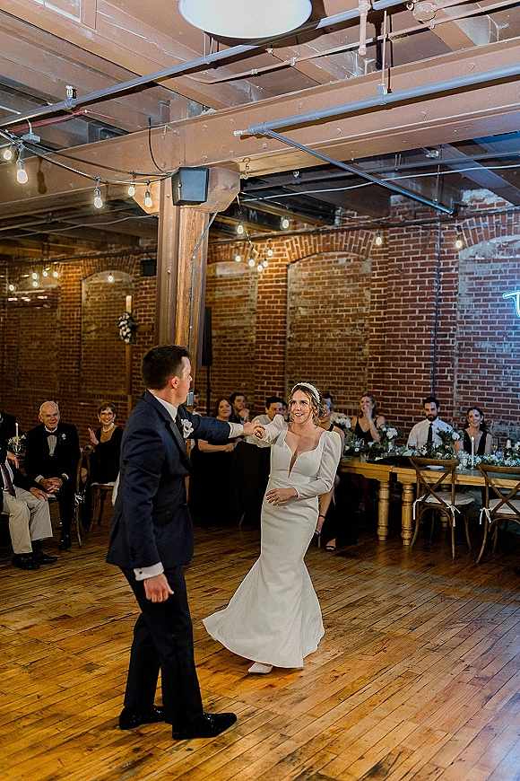 First dance as bride twirls in a long sleeve wedding dress with groom in suit, candlelit head table and string lights behind