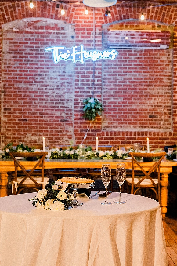 Sweetheart table decor with a neon wedding sign, string lights, greenery garland, candles and champagne flutes against a brick wall backdrop