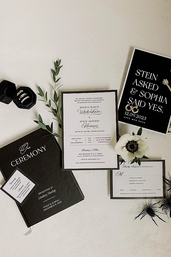Invitation suite with black wedding invitations, save the date, RSVP and program cards beside rings, earrings, greenery, and thistle on a neutral tabletop
