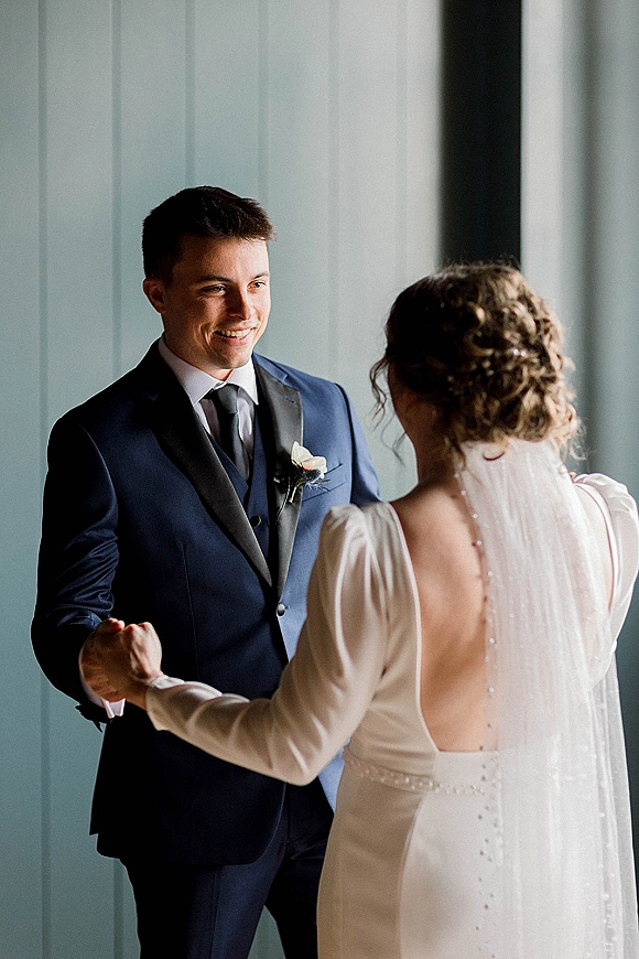 First look moment as bride in long sleeve dress and veil holds hands with groom in blue suit by window light against paneled walls