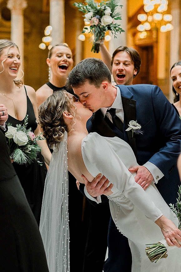 Wedding kiss portrait with bride dipped in groom’s arms as bridesmaids cheer, bouquets raised under warm chandelier lights and columns