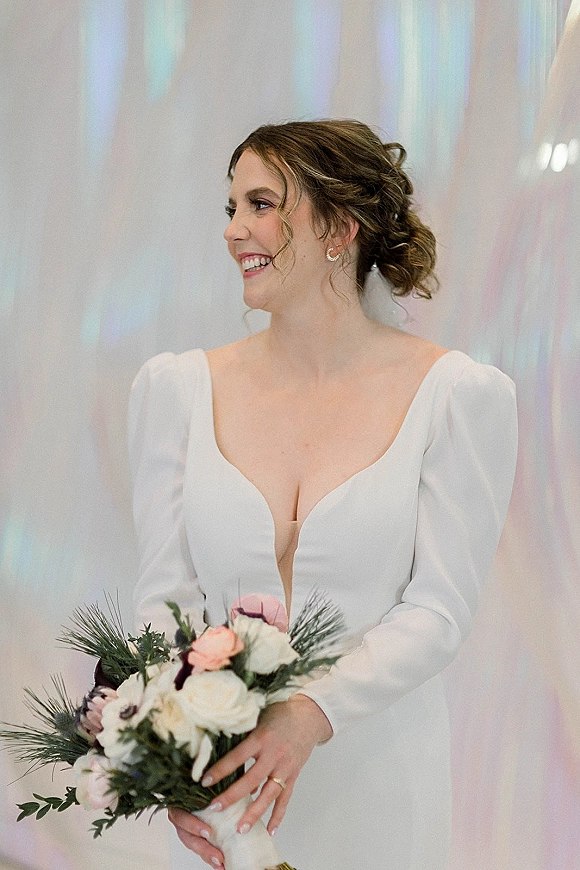 Bridal portrait of a bride holding bouquet, smiling in a white long sleeve deep V gown with earrings against an iridescent backdrop