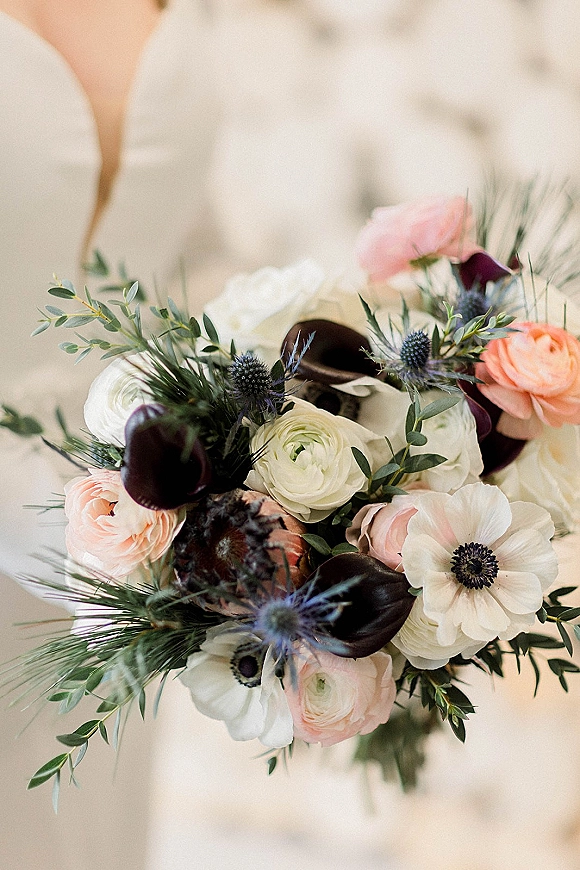 Bridal bouquet with white and blush bouquet tones, featuring peach roses, ranunculus, anemones, and dark calla lilies against a neutral outdoor blur