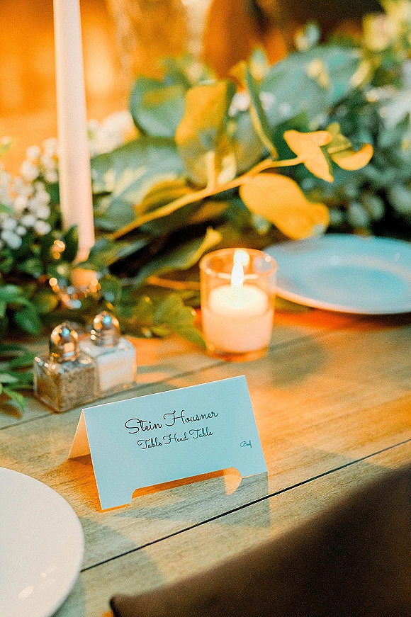 Wedding place card with calligraphy place cards beside a glass votive candle and eucalyptus garland on a rustic wood reception table under warm light