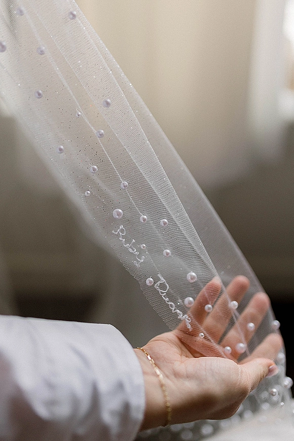 Wedding veil close-up with pearl wedding veil details and embroidered lettering, held beside a bridal robe sleeve against a soft neutral background