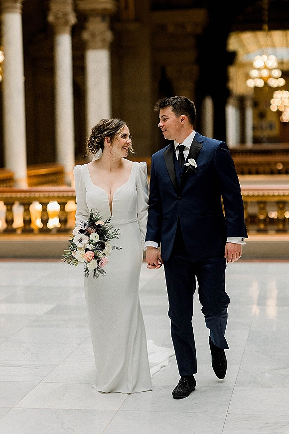 Couple portrait of bride and groom holding hands, bride looking at groom in a grand hall with columns and chandelier lights, holding bouquet