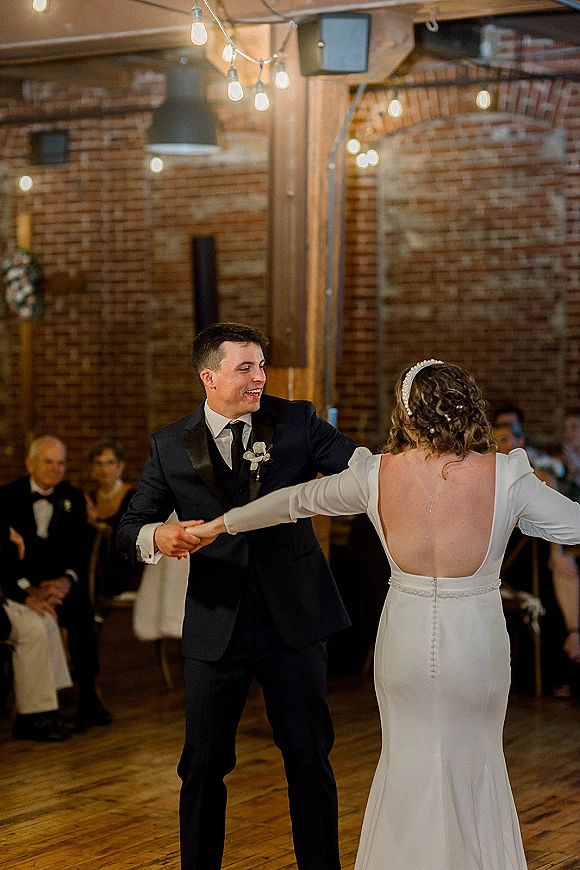 First dance as bride in long sleeve open-back wedding dress and groom in suit spin under string lights before brick wall guests