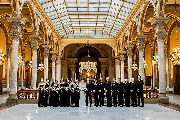 Wedding party portrait with bride and groom centered, bridesmaids in black dresses holding bouquets, groomsmen in tuxedos in a grand marble hall