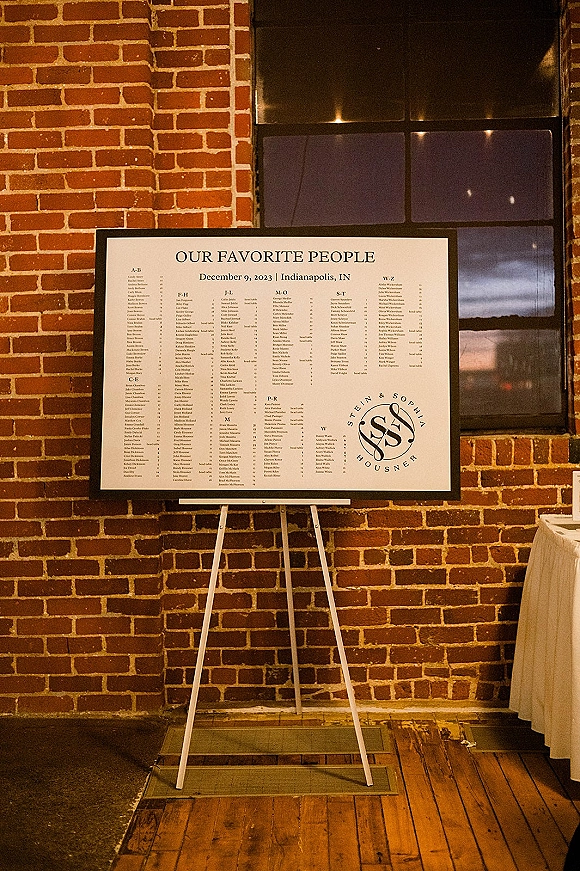 Wedding seating chart sign in a black frame on an easel, featuring calligraphy and monogram logo against a brick wall backdrop