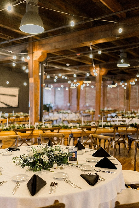 Reception tablescape with round wedding table setup, white linens, black napkins, greenery garland, white flowers and candles beneath string lights