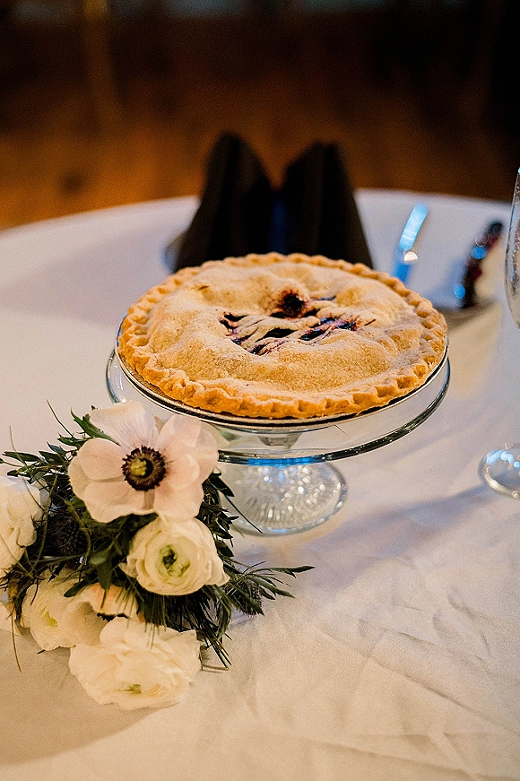 Wedding dessert table featuring a fruit pie on a glass cake stand with white flowers and greenery against a wooden wall backdrop