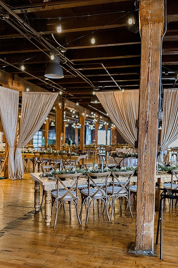 Reception tablescape with farm table reception styling, greenery garlands and place settings under white draping and string lights in a wood-beam venue