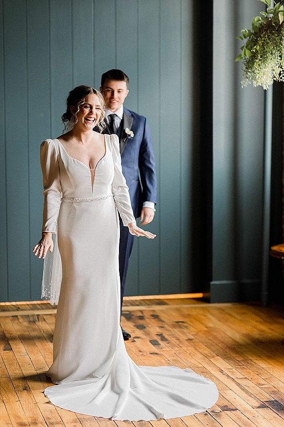 First look moment as bride in long-sleeve deep V wedding dress laughs while groom turns, against dark paneled wall and greenery installation