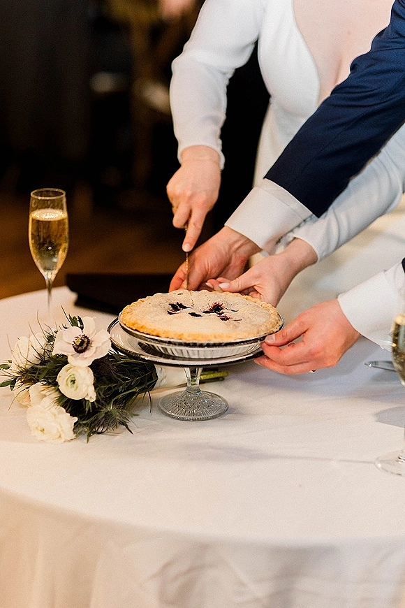 Wedding cake cutting as newlyweds slice a berry pie on a cake stand with knife, champagne flutes, and floral greenery on white tablecloth indoors