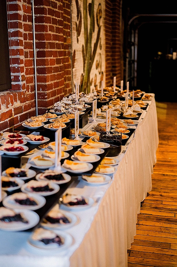 Wedding dessert table with assorted mini pies on tiered stands and labeled place cards, set against a brick wall with mural art