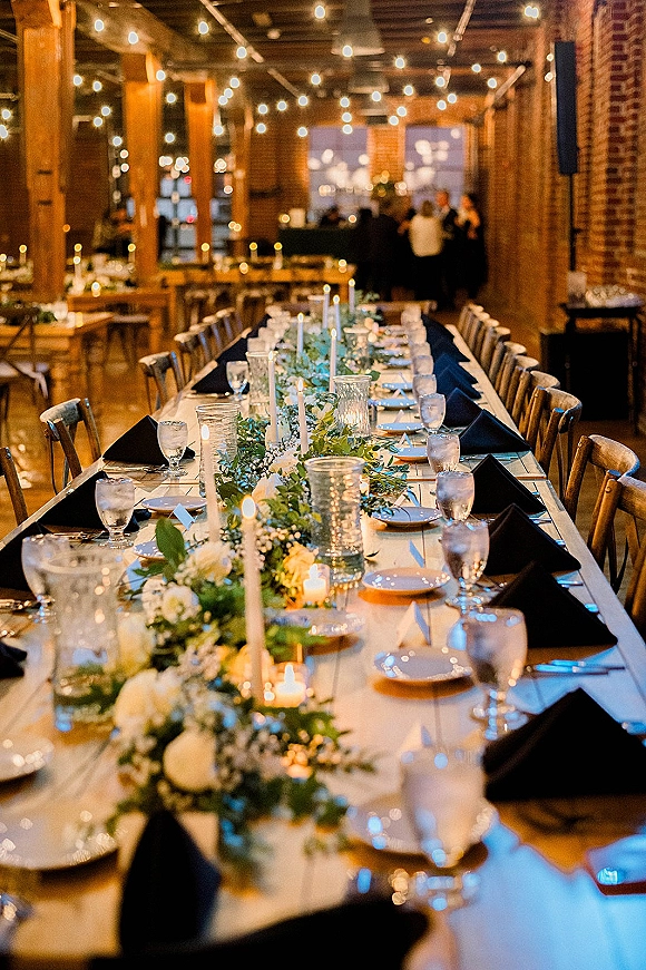 Reception tablescape with long banquet table wedding styling, greenery garland and taper candles amid brick walls, wood beams, and string lights