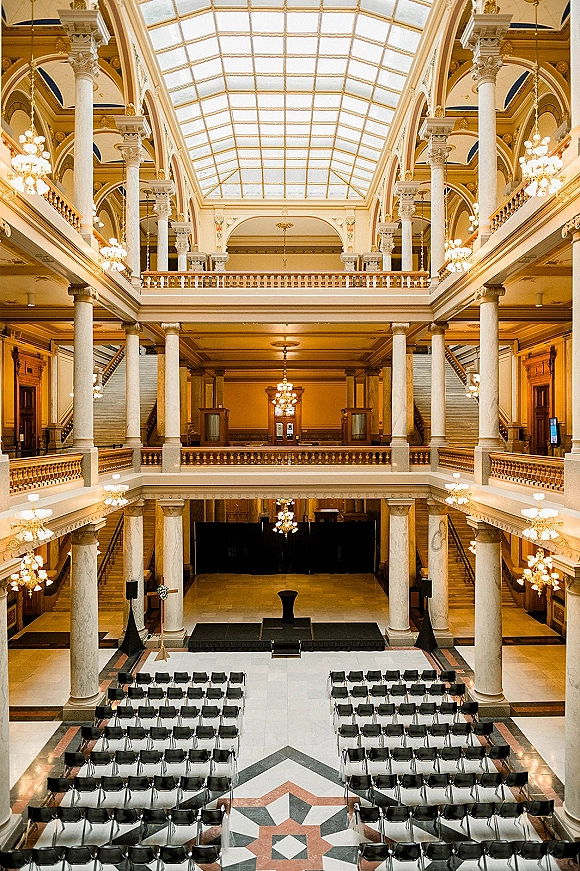 Indoor ceremony setup in a grand atrium wedding ceremony with black chairs facing a stage, marble columns, chandelier, and glass skylight backdrop