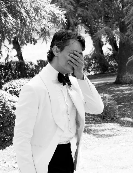 Groom portrait of a groom in a white tuxedo jacket with black bow tie and boutonniere, standing in sunlit garden trees and lawn