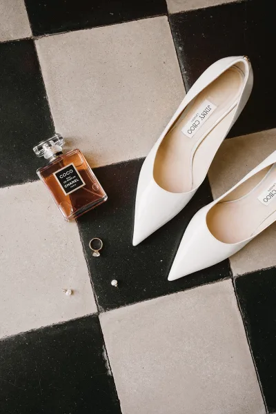 Wedding shoes flatlay with white pointed-toe heels and a perfume bottle, engagement ring, and pearl earrings on checkered tile floor