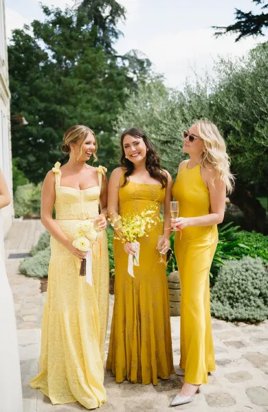 Bridesmaid portrait of three women in yellow bridesmaid dresses holding bouquets, laughing on a stone patio with garden greenery behind